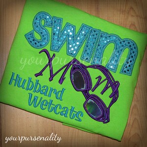 Może przedstawiać: Jasnozielona koszulka z wyhaftowanym napisem "SWIM MOM" dużymi, błyszczącymi niebieskimi literami. Poniżej znajduje się niebieski napis "Hubbard Wetcats" oraz wyhaftowana grafika fioletowych gogli pływackich z odblaskowymi soczewkami.