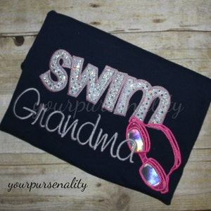 Może przedstawiać: Ciemnogranatowy t-shirt z napisem SWIM wykonanym srebrnymi, brokatowymi literami z r&oacute;żowym obszyciem oraz napisem Grandma wyhaftowanym jasnoszarym pismem. Poniżej wyhaftowano jaskrawor&oacute;żowe okularki pływackie z opalizującymi szkłami.