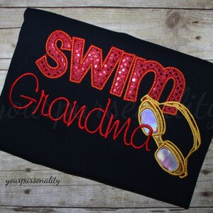 Może przedstawiać: Czarna koszulka z wyhaftowanym czerwonym napisem "Swim Grandma". Słowo "Swim" wypełnione jest czerwonymi cekinami, a "Grandma" napisane jest czerwoną kursywą. Widoczne są r&oacute;wnież wyhaftowane złote okulary pływackie z opalizującymi szkłami.