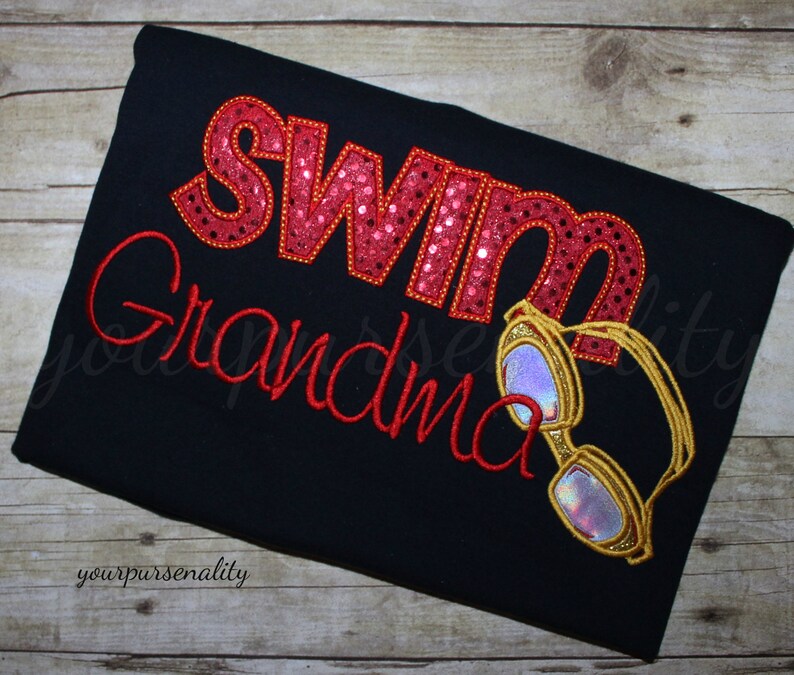 Może przedstawiać: Czarna koszulka z wyhaftowanym czerwonym napisem "Swim Grandma". Słowo "Swim" wypełnione jest czerwonymi cekinami, a "Grandma" napisane jest czerwoną kursywą. Widoczne są r&oacute;wnież wyhaftowane złote okulary pływackie z opalizującymi szkłami.