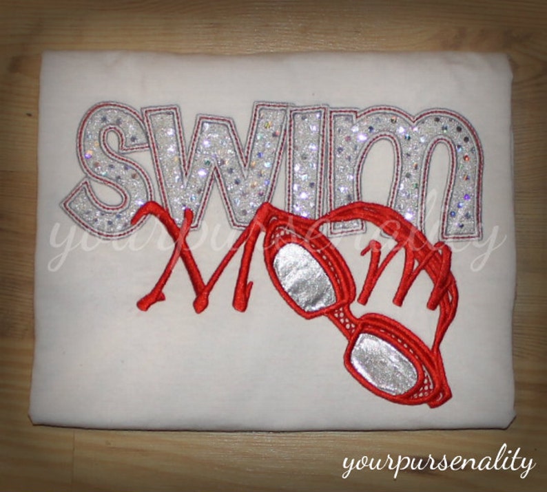 Może przedstawiać: Biały t-shirt z wyhaftowanym napisem SWIM srebrnymi, brokatowymi literami oraz napisem MOM czerwoną kursywą. Czerwone okulary pływackie ze srebrnymi szkłami są umieszczone nad słowem MOM.