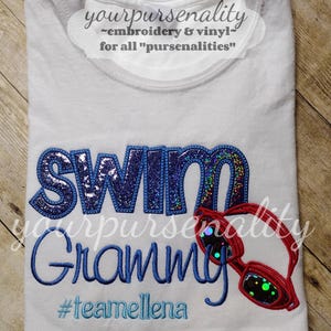 Może przedstawiać: Biały t-shirt z napisem SWIM wykonanym ciemnoniebieskimi, brokatowymi literami, wyhaftowanym niebieskim słowem Grammy oraz czerwonymi okularami pływackimi. Poniżej tekstu wyhaftowano jasnoniebieską nicią hashtag #teamellena.