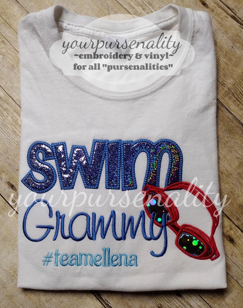 Może przedstawiać: Biały t-shirt z napisem SWIM wykonanym ciemnoniebieskimi, brokatowymi literami, wyhaftowanym niebieskim słowem Grammy oraz czerwonymi okularami pływackimi. Poniżej tekstu wyhaftowano jasnoniebieską nicią hashtag #teamellena.