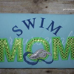 Może przedstawiać: Jasnoniebieski t-shirt z wyhaftowanym napisem SWIM ciemnoniebieskimi literami nad słowem MOM w kolorze limonkowym w czarne kropki. Litera O w słowie MOM została zastąpiona srebrną, brokatową grafiką pływaka z niebieskimi falami.