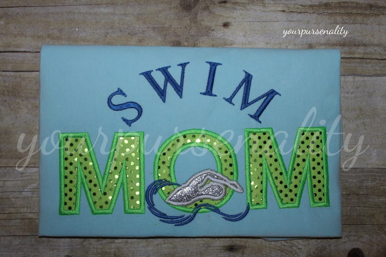 Może przedstawiać: Jasnoniebieski t-shirt z wyhaftowanym napisem SWIM ciemnoniebieskimi literami nad słowem MOM w kolorze limonkowym w czarne kropki. Litera O w słowie MOM została zastąpiona srebrną, brokatową grafiką pływaka z niebieskimi falami.
