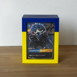 May include: A blue and yellow card storage box with a card featuring the text "Herald of the Arcane." The box is rectangular with a lid, and the card is visible through a window. The box is on a wooden surface.