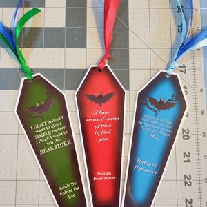 May include: Three coffin-shaped bookmarks with different colored backgrounds and bat illustrations. The green bookmark says "I DON'T believe I want to give a SIMPLE answer. I think I want to tell the REAL STORY. Louis De Pointe Du Lac". The red bookmark says "I have crossed ocean of time to find you. Dracula Bram Stoker". The blue bookmark says "We breathe the light. We breathe the music. We breathe the moment. As it passes through US. Lestat de Lioncourt".