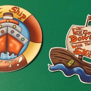 May include: Two colorful stickers with a nautical theme. The first sticker is round and features a red, white, and blue ship with the text "I Ship It!" The second sticker is shaped like a ship and features a brown ship with the text "I Will Go Down With This Ship!"
