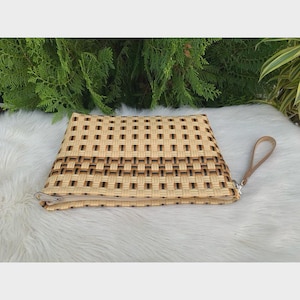 May include: A woven clutch in shades of tan and brown, featuring a rectangular shape with a zipper closure. The clutch has a woven pattern and a small, attached handle. The background is a soft, white surface with green foliage.