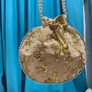 Puede incluir: Un bolso redondo beige con cordón y bordados florales dorados y lentejuelas. El bolso tiene una correa de cuentas y un ribete de cuentas alrededor del borde. El cordón es dorado. El bolso se utiliza probablemente para llevar objetos pequeños.