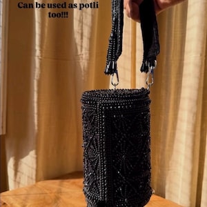 May include: Black beaded cylindrical handbag with a beaded handle and fringe. The text "Can be used as potli too!!!" is visible. The bag is held up by a hand.