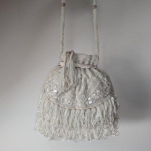May include: A white drawstring handbag featuring intricate beadwork, shimmering sequins, and a long beaded fringe along the bottom edge. The bag includes a matching beaded shoulder strap and a braided drawstring closure with decorative tassels.