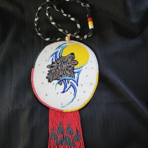 May include: Handmade beaded necklace featuring a central pendant with a howling wolf design against a yellow moon. The pendant has a white background with blue accents and a red beaded fringe. The necklace cord is black and white braided.