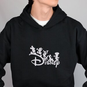 May include: Black hooded sweatshirt featuring a white Disney logo with Mickey Mouse, Donald Duck, Goofy, and Minnie Mouse silhouettes. The sweatshirt has a front pocket and a drawstring hood. The model is wearing blue jeans.