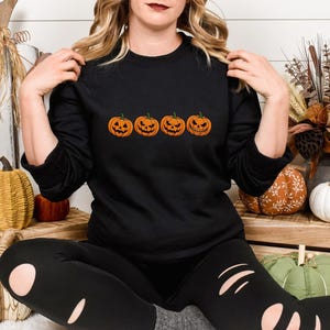 May include: A black long-sleeve sweatshirt featuring a horizontal row of four orange jack-o'-lantern pumpkins with distinct carved facial expressions printed across the center of the chest, paired with black leggings featuring distressed cutouts.