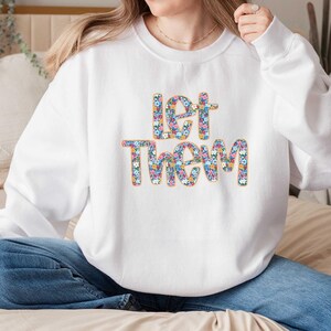 May include: A white crewneck sweatshirt featuring the text "let them" printed in a colorful floral pattern. The letters are filled with small blue, pink, yellow, and white flowers, outlined in a thin orange border against the white fabric.