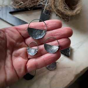 May include: Three silver hoop earrings with a hammered half-circle design. The earrings are different sizes and are displayed on a hand.