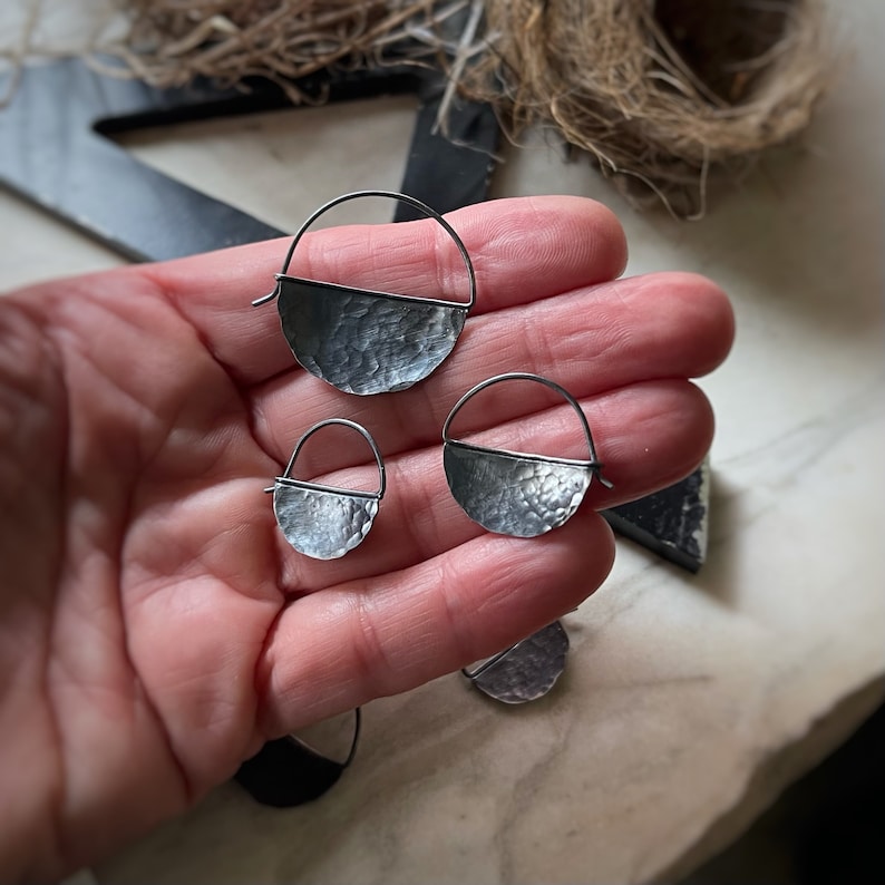 May include: Three silver hoop earrings with a hammered half-circle design. The earrings are different sizes and are displayed on a hand.