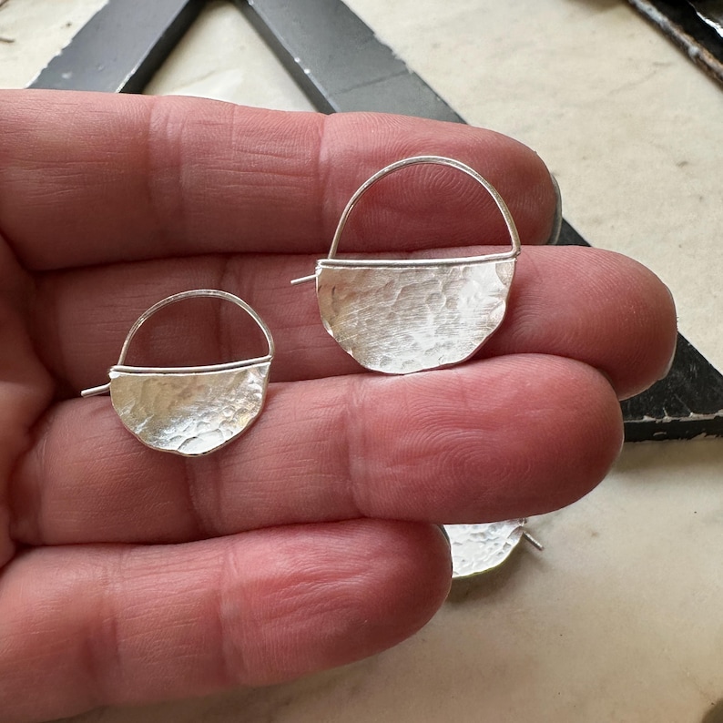 May include: A pair of silver earrings with a hammered, half-circle design. The earrings have a simple wire hoop that goes through the earlobe.