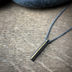 May include: A silver-toned necklace with a rectangular pendant. The pendant is dark gray with a textured gold-colored edge. The necklace chain is also silver-toned and rests on a wooden surface.
