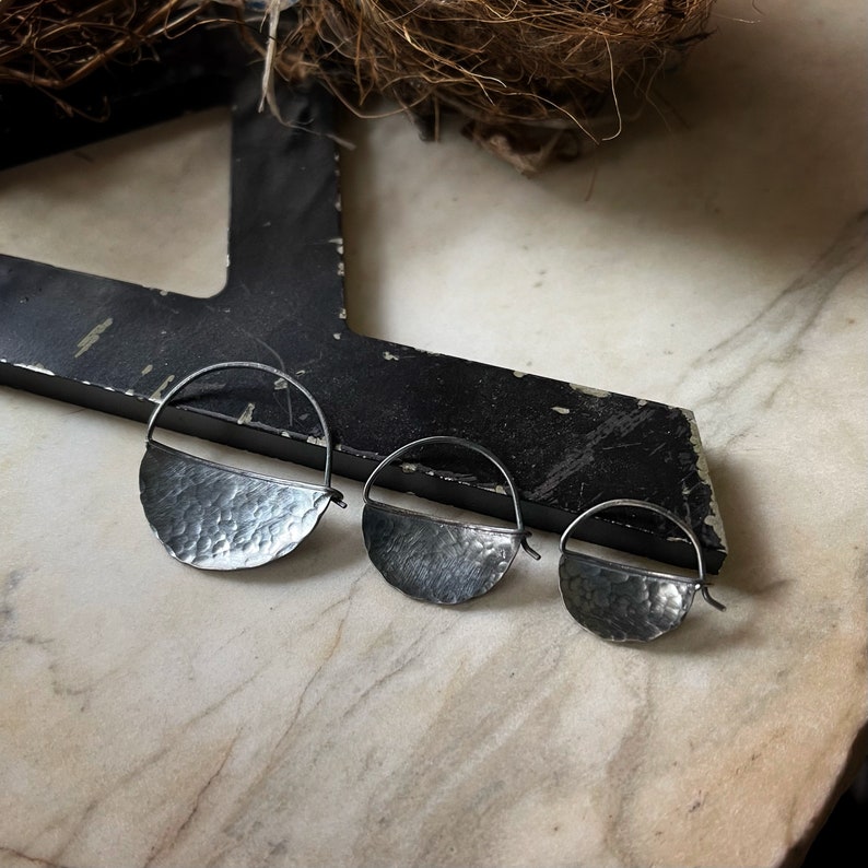 May include: Three silver hoop earrings with a hammered, half-circle design. The earrings are different sizes, with the largest earring on the left and the smallest earring on the right.