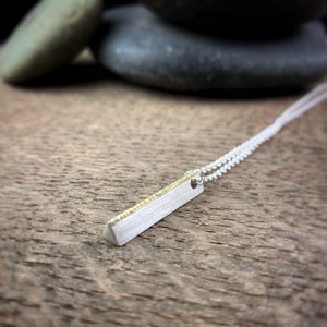 May include: A silver bar pendant necklace with a gold-colored accent. The pendant is rectangular with a brushed finish and hangs from a delicate silver chain. The necklace is displayed on a wooden surface.