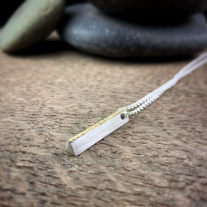 May include: A silver bar pendant necklace with a gold-colored accent. The pendant is rectangular with a brushed finish and hangs from a delicate silver chain. The necklace is displayed on a wooden surface.