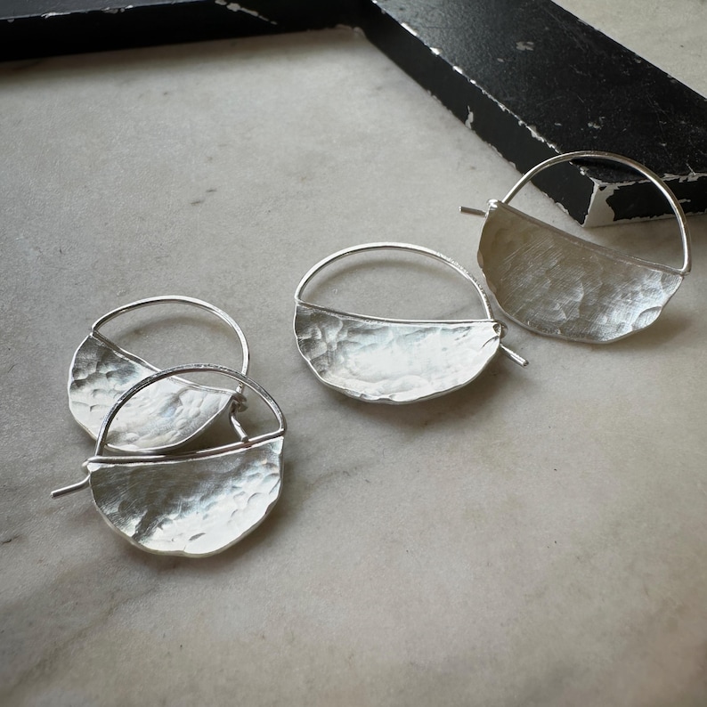 May include: Three pairs of silver hoop earrings with a hammered half-circle design. The earrings are simple and elegant.