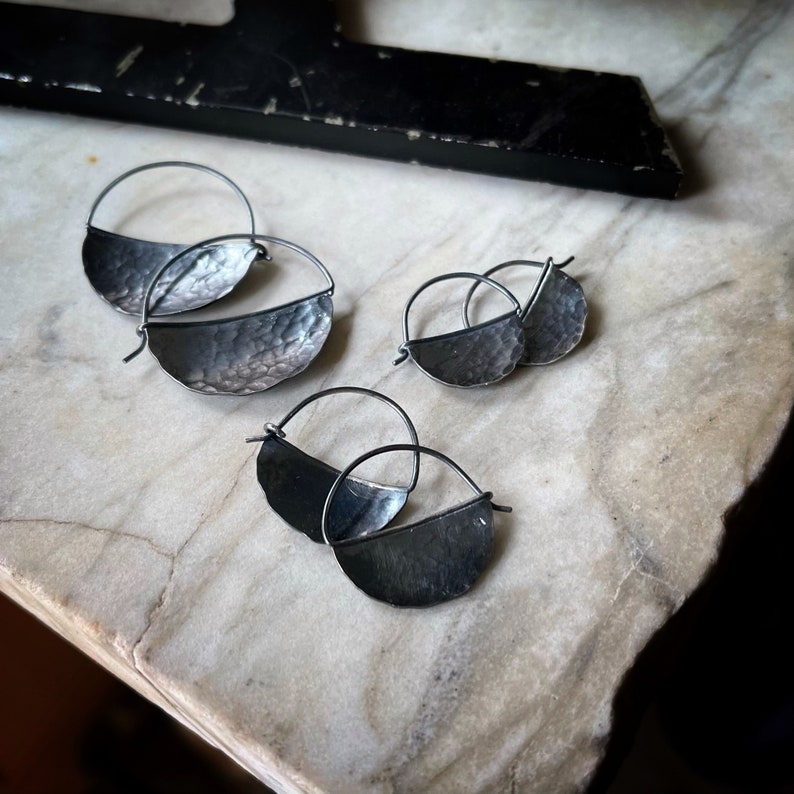 May include: Three pairs of silver hoop earrings with a hammered half-circle design. The earrings are displayed on a white marble surface.