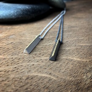 May include: Two silver necklaces with rectangular pendants. The pendants are dark gray with a gold-colored edge and a triangular tip. The necklaces are displayed on a wooden surface.