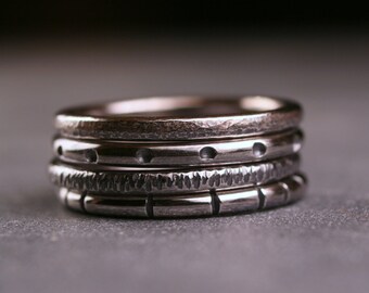 May include: A set of four silver rings stacked on top of each other. The rings have different textures and patterns, including hammered, dotted, and ridged.