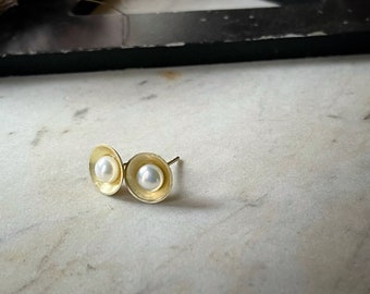 May include: A pair of gold stud earrings with a pearl in the center of each. The earrings are on a white marble surface.