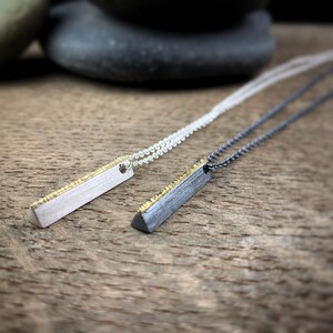 May include: Two minimalist bar pendant necklaces. One is silver-toned with a gold-colored textured edge, the other is dark gray with a gold-colored textured edge. Both hang on delicate chains. The pendants are displayed on a wooden surface.
