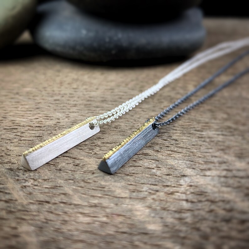 May include: Two minimalist bar pendant necklaces. One is silver-toned with a gold-colored textured edge, the other is dark gray with a gold-colored textured edge. Both hang on delicate chains. The pendants are displayed on a wooden surface.