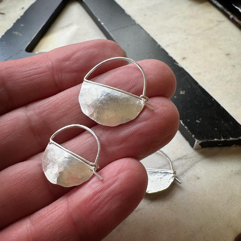 May include: A pair of silver hoop earrings with a hammered half-circle design. The earrings are simple and elegant.