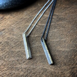 May include: Two silver necklaces with rectangular pendants. One necklace has a silver chain, the other a dark chain. The pendants have a textured gold edge. The necklaces are displayed on a wooden surface.