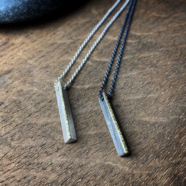 May include: Two silver necklaces with rectangular pendants. One necklace has a silver chain, the other a dark chain. The pendants have a textured gold edge. The necklaces are displayed on a wooden surface.