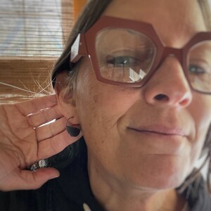 May include: A person wearing black half-circle earrings. The earrings are made of a dark material and have a shiny finish.