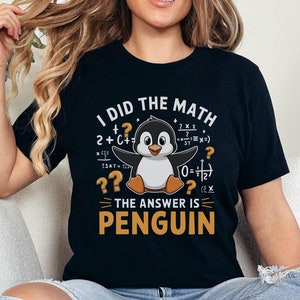 Op de afbeelding: Zwart T-shirt met een witte pinguïn afbeelding en de tekst "Ik heb de wiskunde gedaan, het antwoord is pinguïn". De pinguïn is omringd door wiskundige vergelijkingen.