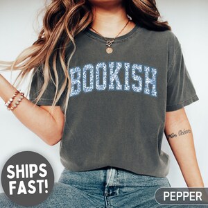 May include: Dark grey t-shirt with the word "BOOKISH" in blue floral print. The shirt is a relaxed fit and made of soft material. The shirt is a comfortable choice for book lovers.