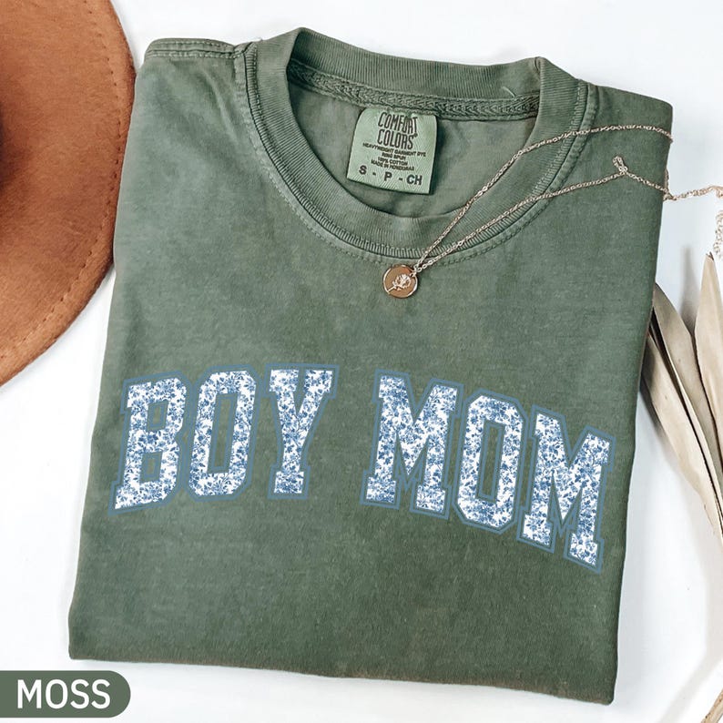 K&ouml;nnte beinhalten: Ein olivgr&uuml;nes T-Shirt mit dem Text "BOY MOM" in einem blauen Blumenmuster. Eine goldene Halskette mit Anh&auml;nger liegt auf dem Shirt. Das Shirt ist gefaltet und auf einer wei&szlig;en Oberfl&auml;che platziert.