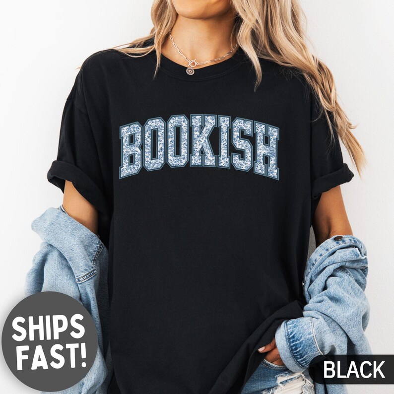 May include: Black t-shirt with the word "BOOKISH" in a blue floral design. The shirt has short sleeves and a crew neck. The text "SHIPS FAST!" is in a white circle on the left side, and the word "BLACK" is on the right side.