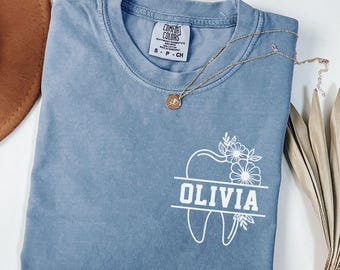 May include: A blue jean colored t-shirt with a white graphic on the chest featuring a tooth outline, floral accents, and the name OLIVIA printed in a bold font. The shirt has a crew neckline and a Comfort Colors brand label inside the collar.