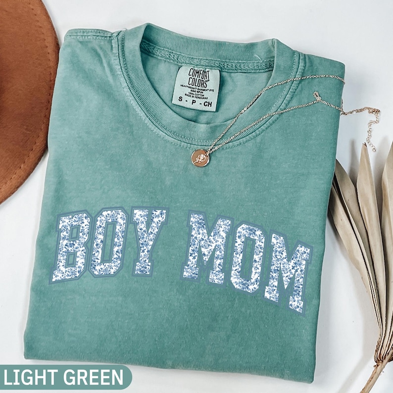 K&ouml;nnte beinhalten: Ein hellgr&uuml;nes T-Shirt mit dem Text "BOY MOM" in einem floralen Design. Das Shirt ist gefaltet und hat ein Comfort Colors-Etikett. Eine goldene Halskette mit Anh&auml;nger liegt auf dem Shirt.