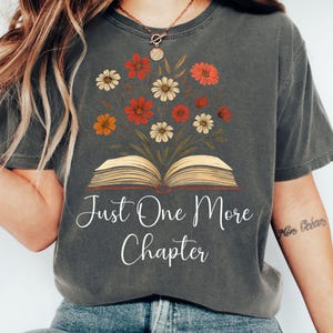 以下が含まれることがあります： フローラルデザインと「Just One More Chapter」のテキストが入った濃いグレーのTシャツ。グラフィックは、赤、オレンジ、クリーム色の花が咲く開いた本が特徴です。本好きのためのカジュアルなTシャツ。