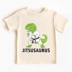 May include: Cream-colored t-shirt featuring a green cartoon dinosaur in a white karate uniform with a black belt. The text "Jitsusaurus" is printed in black below the dinosaur. The shirt is made of soft material.