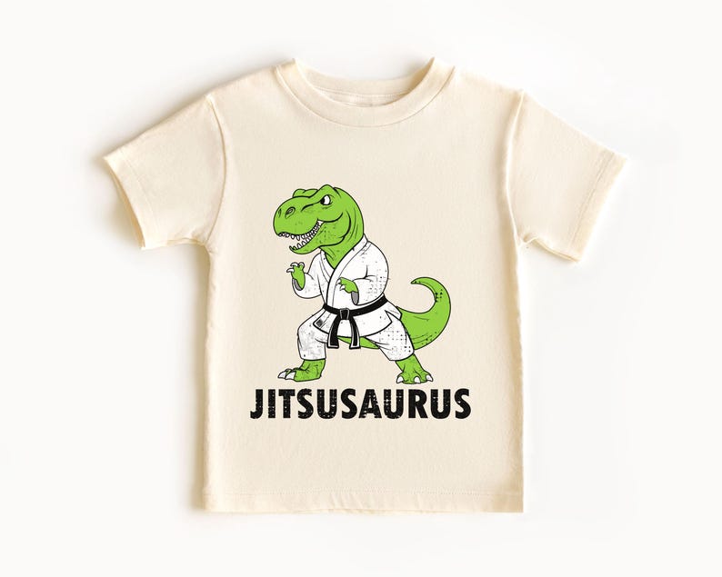 May include: Cream-colored t-shirt featuring a green cartoon dinosaur in a white karate uniform with a black belt. The text "Jitsusaurus" is printed in black below the dinosaur. The shirt is made of soft material.