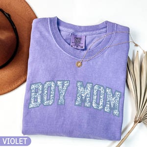 K&ouml;nnte beinhalten: Ein violettes Comfort Colors T-Shirt mit dem Text "BOY MOM" in einem floralen Design. Eine goldene Halskette mit Anh&auml;nger liegt auf dem Shirt. Ein brauner Hut und ein getrocknetes Palmblatt sind im Hintergrund zu sehen.