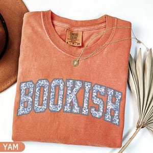 May include: A coral-colored t-shirt with the word "BOOKISH" in a blue floral design. The shirt has a crew neck and short sleeves. A gold necklace with a pendant is draped across the shirt. The shirt is folded on a white surface.