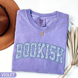 May include: A violet-colored t-shirt with the word "BOOKISH" in a floral design across the front. A gold necklace with a pendant is draped over the shirt. A brown hat and a dried palm leaf are in the background.